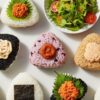 様々な種類のコンビニおにぎりと、小さなサラダや枝豆などのヘルシーな副菜が並べられた、明るく魅力的な俯瞰ショット。バランスの取れた食事を提案している。