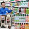 セブンイレブンのドリンクコーナーで、笑顔の日本人男性店長が三ツ矢サイダーのペットボトルを手に持ち、ドリンクの品揃えを説明している様子。