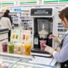 セブンイレブンの店内で、エプロンを着用した日本人女性店長がセブンカフェスムージーマシンを操作し、スムージーを準備している様子。カウンターには複数のスムージーが並べられている。 画像2