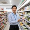 現役の日本人コンビニ店長が、セブンイレブンの冷蔵棚に並んだきゅうりの一本漬けを指し示し、笑顔で歓迎している様子。