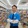 コンビニエンスストアの店長らしき日本人男性が、笑顔で顧客を迎えている。店内は清潔で、セブンイレブンのロゴが見える。