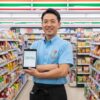 セブンイレブンの制服を着た日本人男性の店長が、店内でタブレットを持ち笑顔で立っている。タブレットには「マイローカルコンビニ」のロゴが表示されている。
