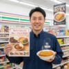 セブンイレブンの制服を着た日本人男性の店長が、カレーフェス2025のポスターとカツカレーを持ってコンビニ店内で笑顔で立っている。