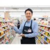 ンビニエンスストアの店内で、笑顔で両手を広げて案内する日本人男性の店長。エプロンを着用し、名札を付けている。