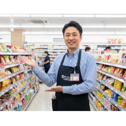 ンビニエンスストアの店内で、笑顔で両手を広げて案内する日本人男性の店長。エプロンを着用し、名札を付けている。