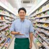 現役の日本人コンビニ店長である齋藤正志が、店内の通路で微笑みながら手招きしている様子。棚には様々なおにぎりや商品が並んでいる。