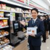 セブンイレブンの店長らしき日本人男性が、店内のセブンカフェ紅茶マシンの前でタブレットを持ち、笑顔で記事を紹介している様子。