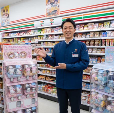 笑顔でコンビニの「ガシャポンボックス」を紹介する日本人男性のコンビニ店長。
