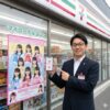 コンビニエンスストアの前に立ち、超ときめき宣伝部のコラボポスターとシェアプリントアプリを表示したスマートフォンを指し示す日本人男性の店長。