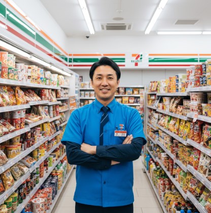 コンビニエンスストアの店長らしき日本人男性が、笑顔で顧客を迎えている。店内は清潔で、セブンイレブンのロゴが見える。