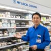 コンビニエンスストアの冷蔵棚の前で、笑顔で商品を紹介する日本人男性の店長。胸元には「齋藤 正志」と書かれた名札が見える。