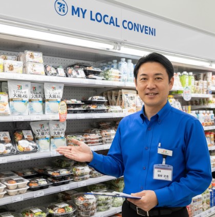 コンビニエンスストアの冷蔵棚の前で、笑顔で商品を紹介する日本人男性の店長。胸元には「齋藤 正志」と書かれた名札が見える。