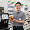 コンビニの制服を着た日本人男性の店長が、セブンカフェのティーマシンとアイスティーのカップを手に笑顔で立っている。背景にはコンビニの店内が広がる。