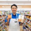 現役の日本人コンビニ店長が、セブンイレブンの店内で「シュガーバターの木」の限定フレーバーとギフトボックスを手に持ち、笑顔で紹介している。