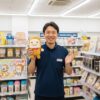 コンビニ店長の日本人男性が店内で「パンどろぼう」のぬいぐるみを持ち、笑顔で手招きしている。背後には雑誌やキャラクターグッズが並んでいる。