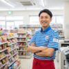 コンビニの制服を着た日本人男性店長が、明るい店内で笑顔で迎えている様子。