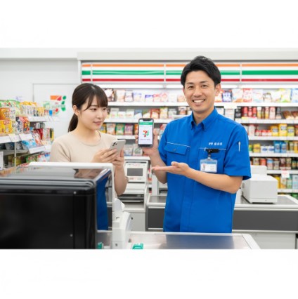 笑顔のセブンイレブン店長が、チケット発券で困っている日本人女性客にスマートフォンの画面を見せながら親切に案内している様子。