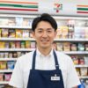 コンビニエンスストアの制服を着た日本人男性店長が、温かい笑顔で挨拶している様子。背景には清潔で整理されたコンビニの店内が広がる。