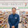 コンビニエンスストアの制服を着て笑顔で立つ日本人男性の店長。背景にはセブンイレブンの商品棚が並び、「マイローカルコンビニ」という看板が見える。