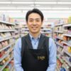 コンビニエンスストアの通路に立つ笑顔の日本人男性店長。黒いエプロンを着用し、親しみやすい雰囲気で読者を出迎えている。