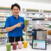 セブンイレブンの店長が、店内で作られた4種類のスムージーとスムージーマシンを指し示している。テーブルにはタブレットでスムージーの栄養成分表が表示されている。