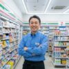 腕を組んで笑顔で立つ日本のコンビニ店長、齋藤正志。店長の制服を着ており、明るく清潔なコンビニエンスストアの通路に立っている。