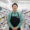 コンビニの制服を着た30代から40代の日本人男性店長が、明るい店内の日用品売り場の前で穏やかに微笑んでいる様子。