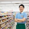 コンビニエンスストアの制服を着て笑顔で立つ、30代くらいの日本人男性店長。背景には清潔感のあるコンビニの商品棚がぼんやりと見える。