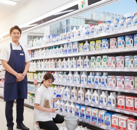 コンビニエンスストアの店内で、笑顔で来店客を迎える日本人男性店長。エプロンを着用し、清潔感のある身だしなみ。