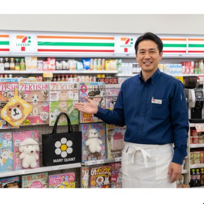 コンビニエンスストアの雑誌コーナーで、さまざまな付録付き雑誌を紹介する日本人男性の店長。ゼクシィやMARY QUANTなどの人気雑誌が陳列され、可愛らしい付録(プーさんの鍋つかみ、MARY QUANTのバッグ、スヌーピーのぬいぐるみなど)が目を引く。店長は清潔なエプロンを着用し、笑顔で手招きしている。