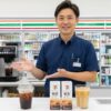 セブンイレブンの制服を着た日本人男性の店長が、セブンカフェのアイスコーヒーとセブンプレミアムの濃縮コーヒーパック、カフェオレを指差して説明している様子。