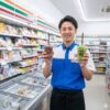 セブンイレブンの制服を着た日本人男性の店長が、店内でフラッペのチョコレート味と抹茶味を笑顔で両手に持ち、読者に商品の魅力をアピールしている様子。背景には商品の並んだ棚と冷凍ケースが写っている。