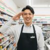 コンビニのエプロンを着用し、カウンター内から笑顔で敬礼する現役店長の日本人男性。