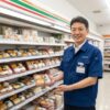 現役の日本人コンビニ店長がセブンイレブンの弁当売り場で幕の内弁当を手に持ち、笑顔で紹介している。