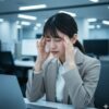 突然の激しい頭痛に苦しみ、オフィスでこめかみを押さえている日本人女性の画像。緊急で薬を探す状況を表現。