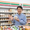 笑顔でセブンイレブンのカップデリ「ピリ辛きゅうり」を持つ現役コンビニ店長の日本人男性