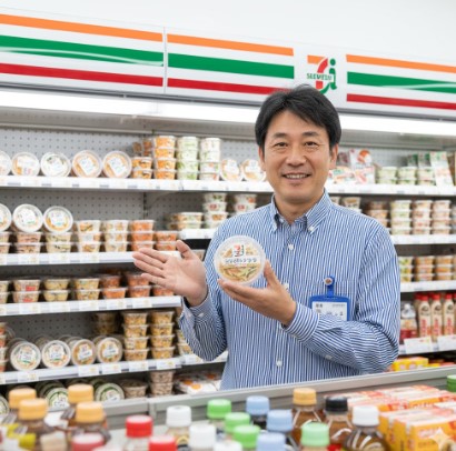 笑顔でセブンイレブンのカップデリ「ピリ辛きゅうり」を持つ現役コンビニ店長の日本人男性