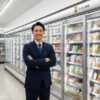 現役の日本人コンビニ店長が、商品の裏側について自信を持って解説する様子。清潔な店舗と冷蔵棚が背景。