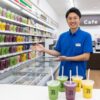 セブンイレブンの店長が、スムージーが並ぶ冷凍ケースの前で笑顔で説明している。手前には完成した様々なスムージーが並び、店内奥にはセブンカフェの機械が見える。