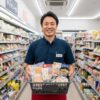 笑顔でコンビニの商品カゴを持つ日本人男性の店長。店内通路を背景にしている。
