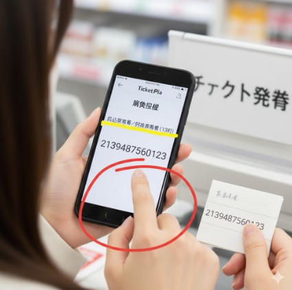 日本人女性がスマートフォンに表示されたチケットぴあの「Myチケット」画面と、手書きメモに書かれた13桁の番号を店員に見せているクローズアップ。