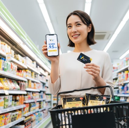 コンビニ店内で、VポイントPayの9.5%還元を示すスマートフォン画面とカードを掲げ、買い物かごに「金のハンバーグ」を入れた笑顔のアジア人女性。