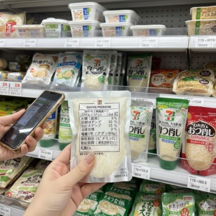 コンビニの冷蔵棚の前で、一人の女性がスマートフォンを片手にセブンプレミアムの大根おろしパッケージ裏の原材料表示を確認している。