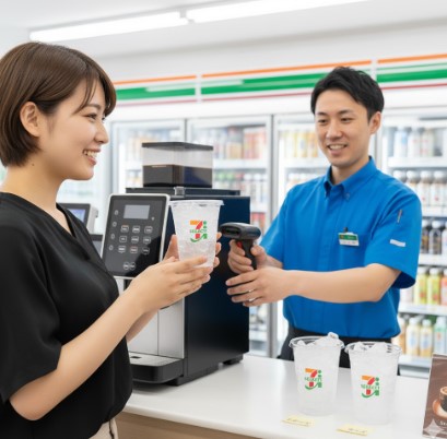 セブンイレブンのレジで、店員が日本人女性客に氷がたっぷり入った専用のプラスチック製アイスコーヒーカップを手渡している。女性客は笑顔でカップを受け取っている。