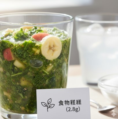 グラスに入ったグリーンスムージーのアップ。ケール、リンゴ、バナナのカットフルーツが見える。手前には「食物繊維(2.8g)」と書かれたカードが置かれ、奥には水と白い粉（砂糖を連想させる）が入ったグラスとボウルがある。