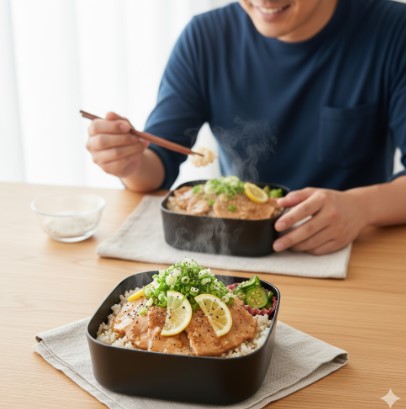 木目のテーブルに置かれた黒い弁当箱に入った、できたてのねぎ塩豚カルビ丼。レモンと刻みネギが乗っており、湯気が立ち上っている。奥には、箸で丼を食べようとしているアジア人男性の笑顔が見える。