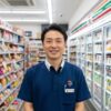 コンビニエンスストアの制服を着て笑顔で立つ日本人男性の店長が、商品棚に囲まれた店内で撮影されている。
